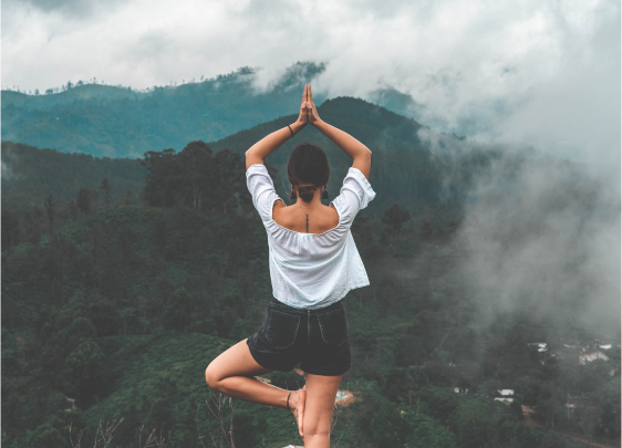 Empowering Yoga Amidst Nature: A woman in a yoga pose, facing a forest with arms aimed upwards, symbolizing the journey to self-discovery with The Joyful Mindset.