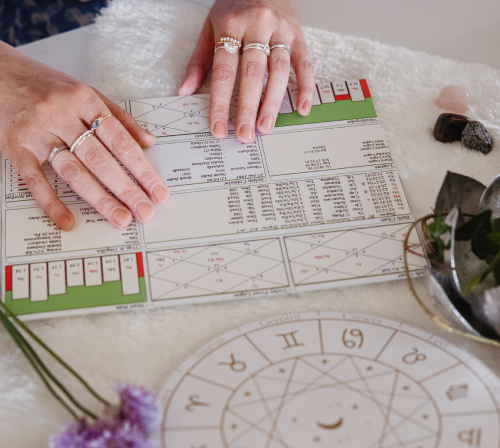 An astrologer's hand drawing a natal chart on paper, surrounded by various astrological charts and tools on a table, with an illustration of the sun and a constellation in the background.