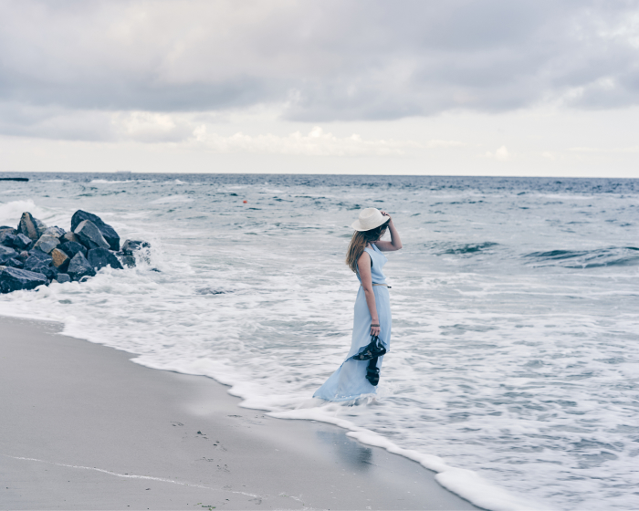 Woman with her back turned, standing in the ocean, wearing a flowing white dress, gazing at the horizon.