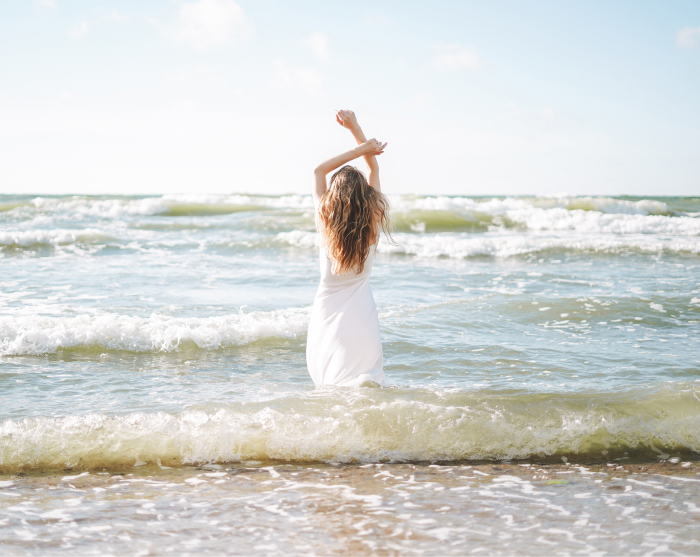 Woman standing in the ocean with her back turned, wearing a beautiful white dress, facing the horizon.